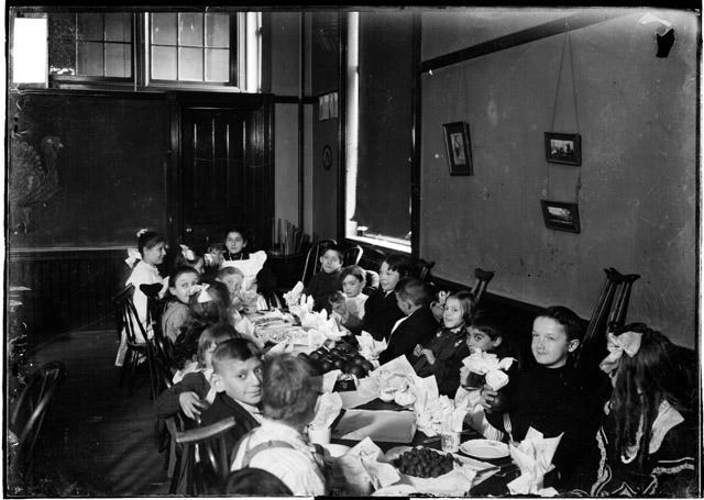image of children with disabilities sitting at a table in a classroom in Chicago, Illinois. The children are elementary age and white, wearing sailor suits, dresses, and suspenders. They look around the room, talking. One boy looks into the camera with a small smile. 